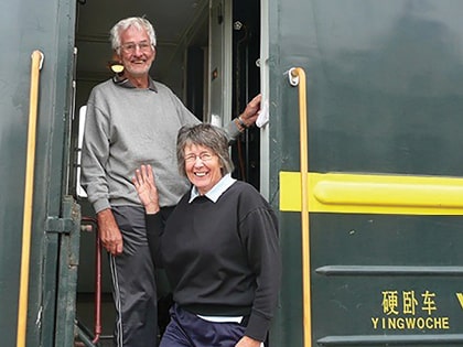 An older man and woman smiling and standing at the entrance of a train. The woman is slightly in front, waving. The train has Chinese characters on its side. Both are casually dressed in sweaters and pants.