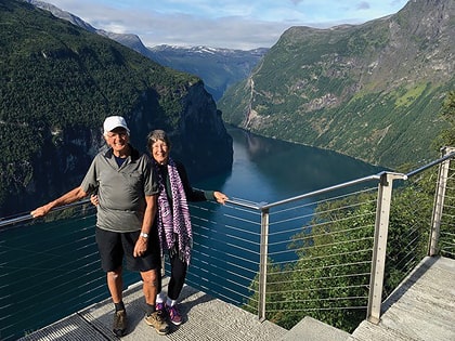 An older couple stands smiling on a scenic overlook with a breathtaking view of a deep blue fjord surrounded by lush, green mountains. They are holding hands, and the sky is partly cloudy.