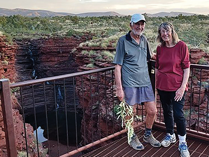 An elderly couple stands on a metal platform overlooking a scenic gorge with red rock walls and greenery. The man holds a small plant, and both are dressed casually. The sky is clear, indicating a bright day.