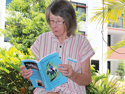 An older woman with glasses reads a blue book while sitting outdoors among lush green plants. She is wearing a striped shirt and appears focused on the book, with a building visible in the background.