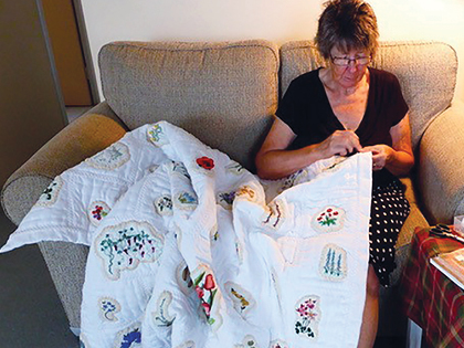 A person sitting on a beige couch is focused on sewing a large white quilt with intricate embroidered designs. They wear glasses and a dark top, surrounded by a cozy indoor setting.