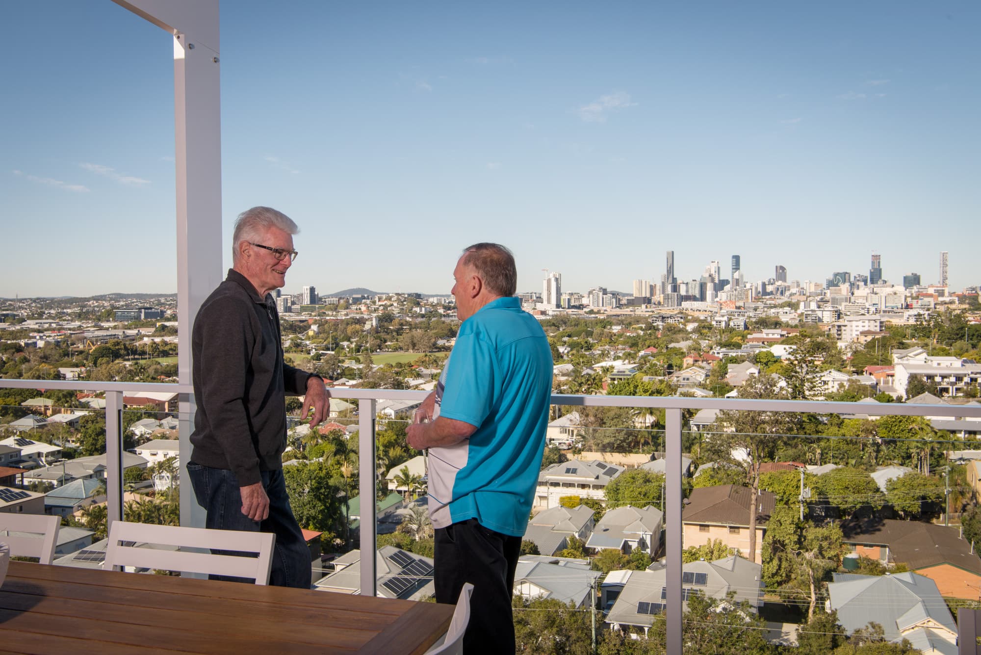 Two men stand on a balcony with a view of a sprawling cityscape under a clear blue sky. The foreground shows outdoor furniture, and the background features a mix of houses and skyscrapers.