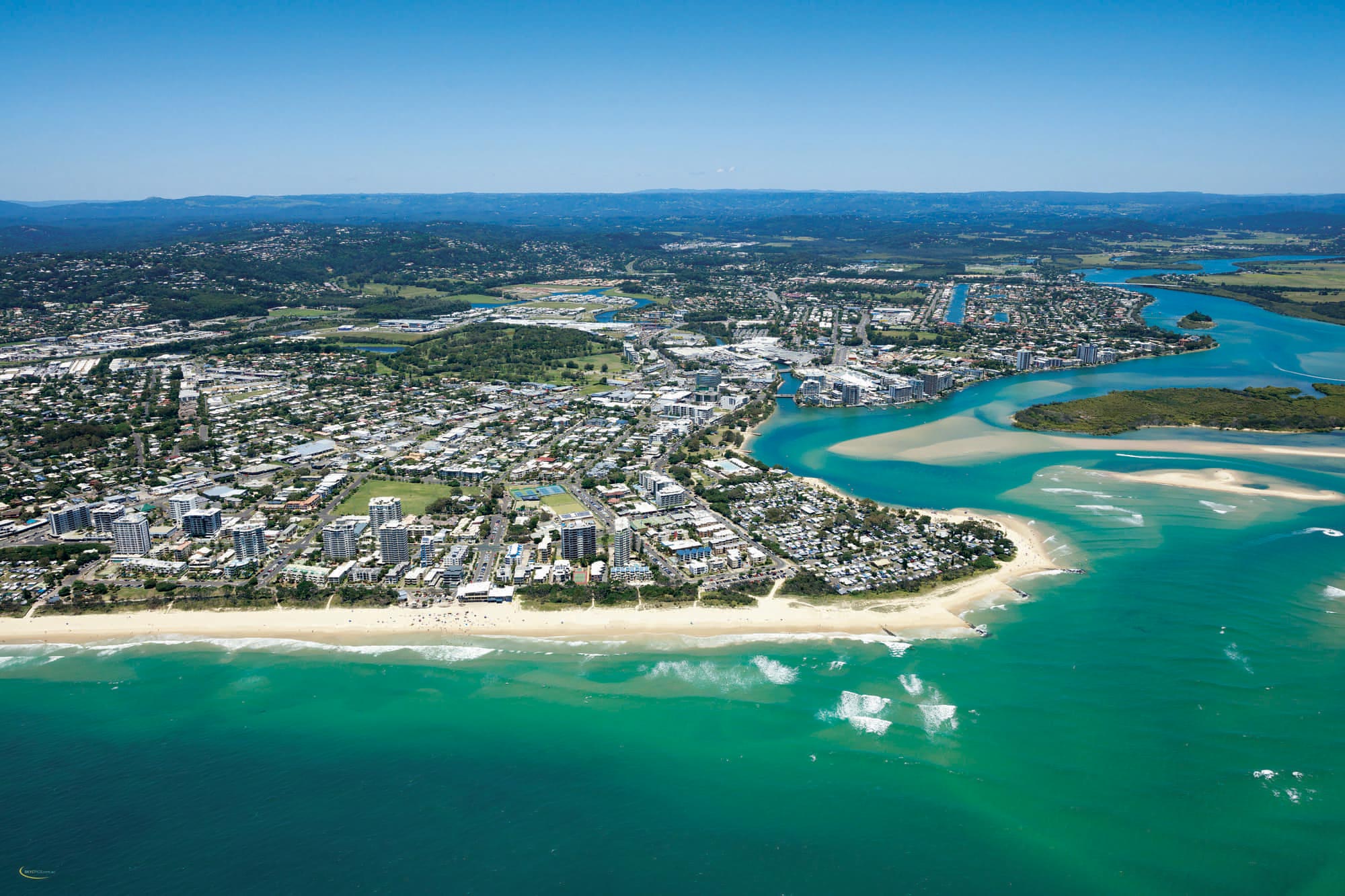 Aerial view of a coastal city with sandy beaches, turquoise ocean, and a river estuary. The cityscape includes residential areas and high-rise buildings under a clear blue sky. Lush greenery surrounds the urban landscape.