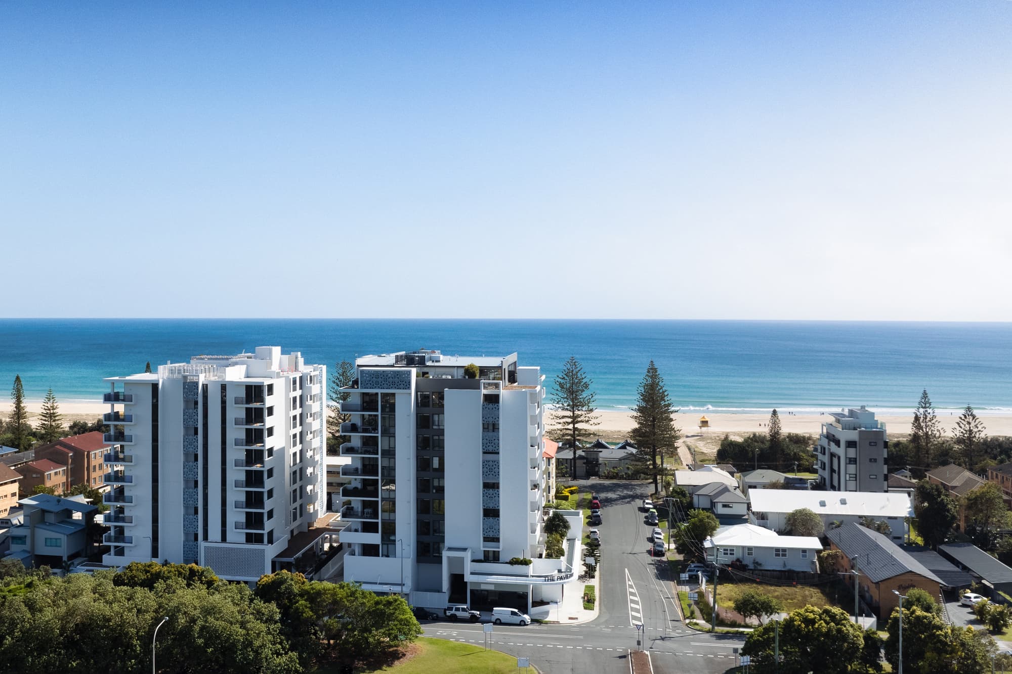 Aerial view of modern beachside apartments with a clear blue sky and ocean in the background. The buildings are surrounded by green trees and residential houses, with a sandy beach visible beyond.