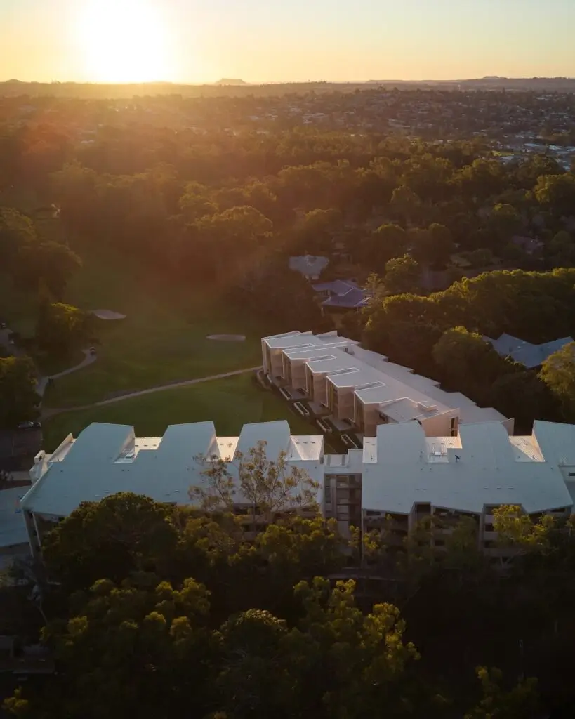 Aerial view of a modern white building at sunrise surrounded by trees and green space, part of the Toowoomba Retirement Community, with the sun low on the horizon and the cityscape visible in the background.