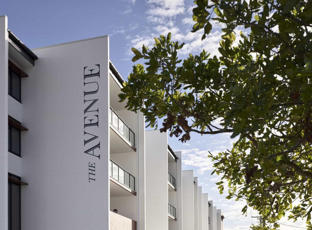 A modern apartment building named "The Avenue" with white walls and glass balconies is partially obscured by tree branches in the foreground. The sky is blue with scattered clouds.