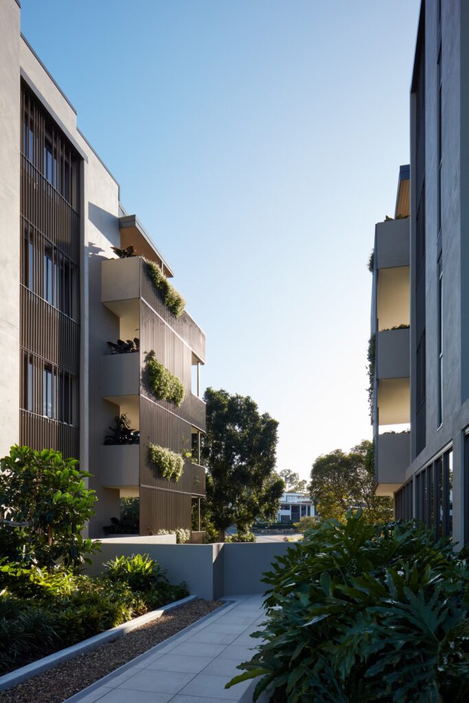 A modern, sunlit apartment complex with plants adorning balconies and greenery lining a walkway. The blue sky and distant trees create a peaceful, inviting atmosphere between two buildings.