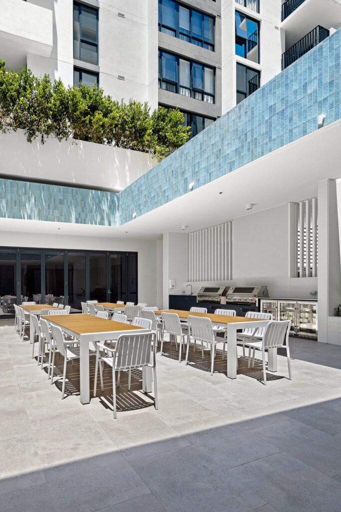 Outdoor dining area with several wooden tables and white chairs on a light stone patio. Modern building with blue-tiled accent wall and greenery above. Large windows and outdoor kitchen area in the background. Bright, sunny day.