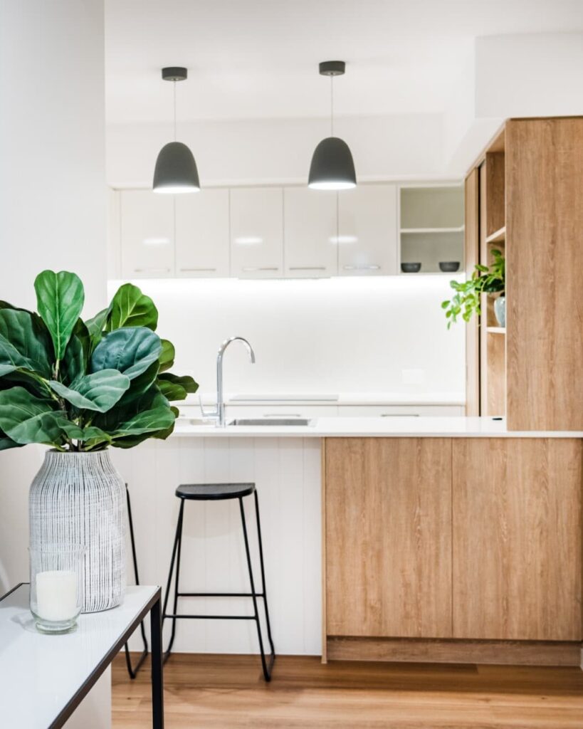 A modern kitchen with a wooden island and white countertops. There are two black barstools, hanging pendant lights, and plants placed on surfaces. The cabinetry is light wood, and a vase with green foliage is on the counter.