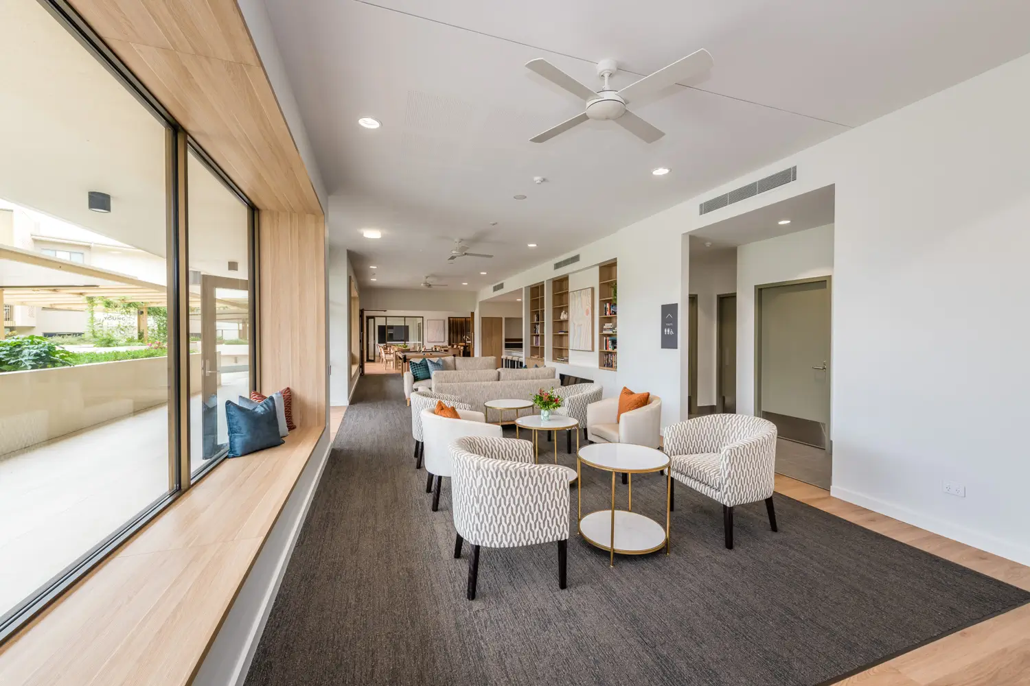 A modern lounge at Toowoomba Retirement Community features round tables and cushioned chairs, a long window seat with pillows, built-in bookshelves, neutral decor, and large windows filling the spacious room with natural light.