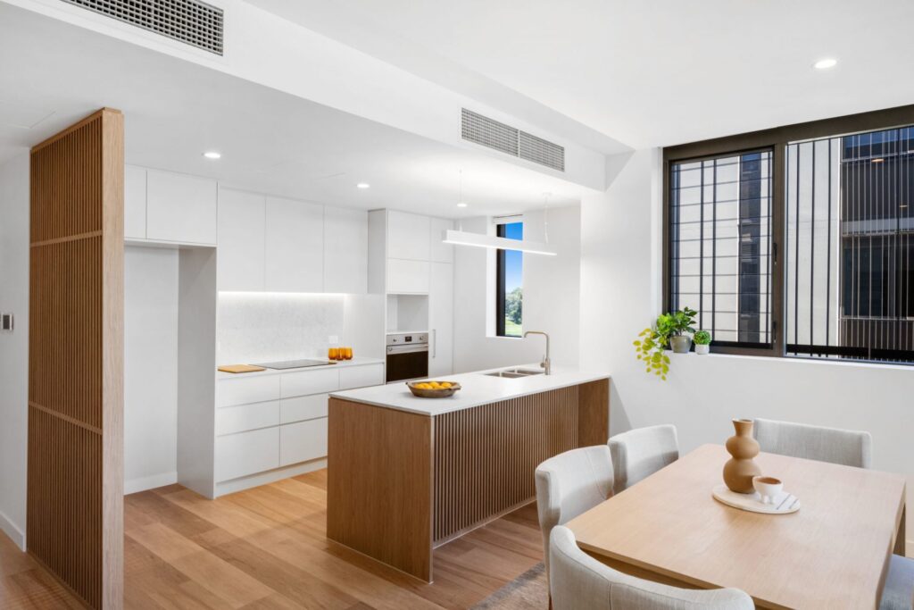 Modern kitchen and dining area in West Brisbane Retirement Community with white cabinets, wood accents, an island sink, a bowl of fruit, and a dining table for four. Large window fills the space with light; plant decor brightens the windowsill.