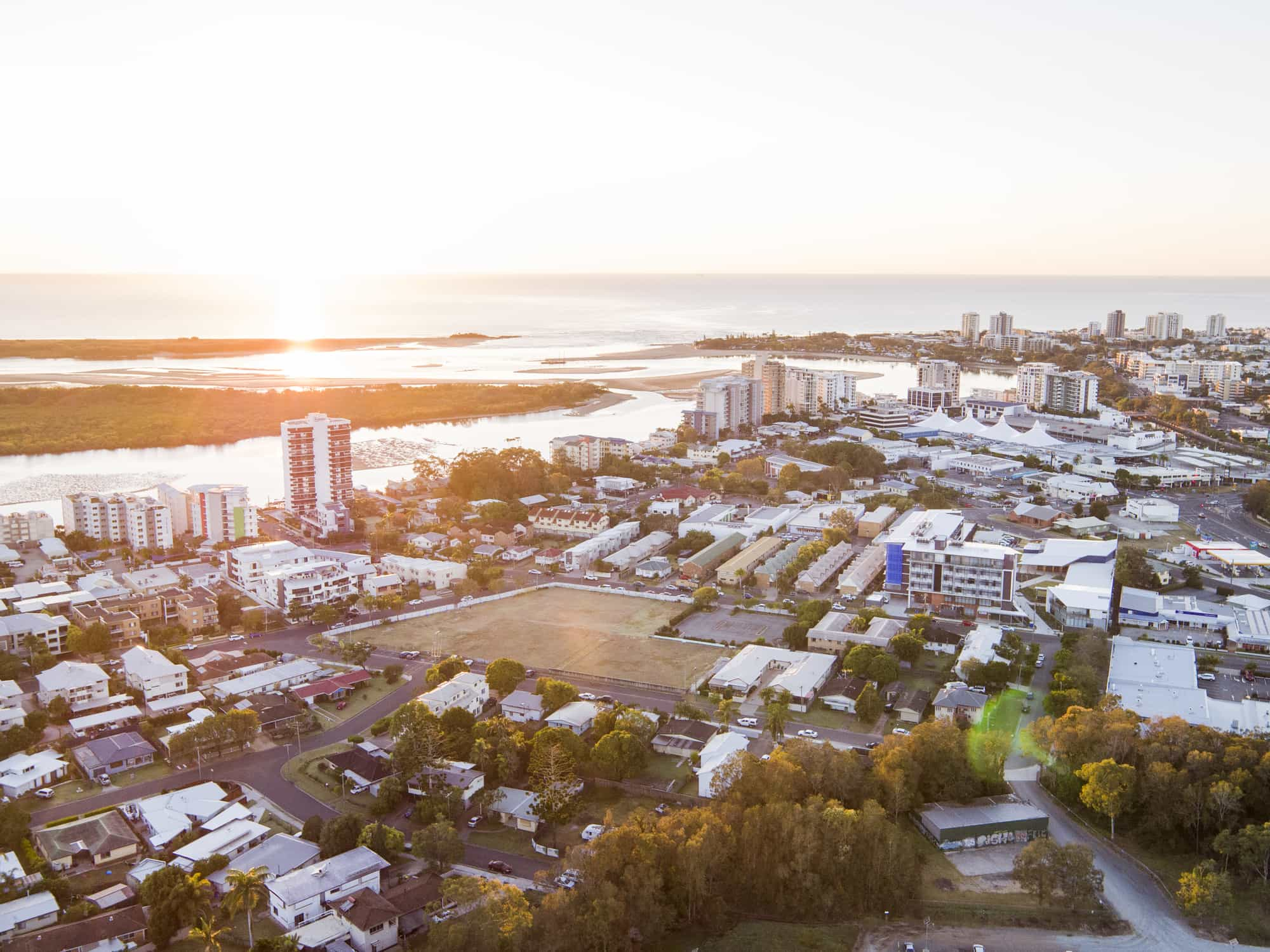 Aerial view of a coastal cityscape at sunset, featuring residential and commercial buildings, lush greenery, and a river flowing into the ocean. The horizon is illuminated by the setting sun, adding a warm, golden hue to the scene.