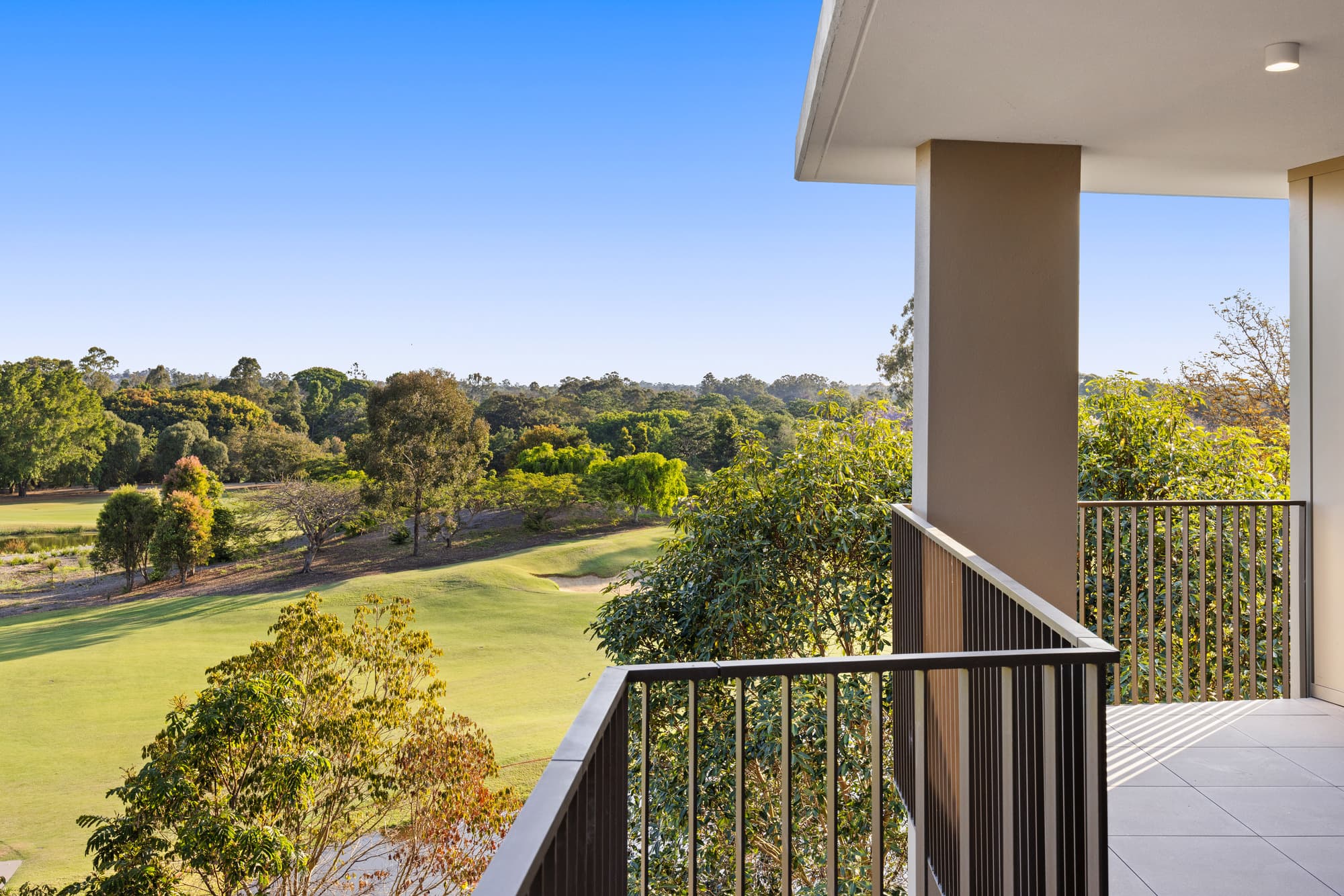 View from a balcony overlooking a lush green golf course surrounded by trees under a clear blue sky, with part of the building and railing visible in the foreground.