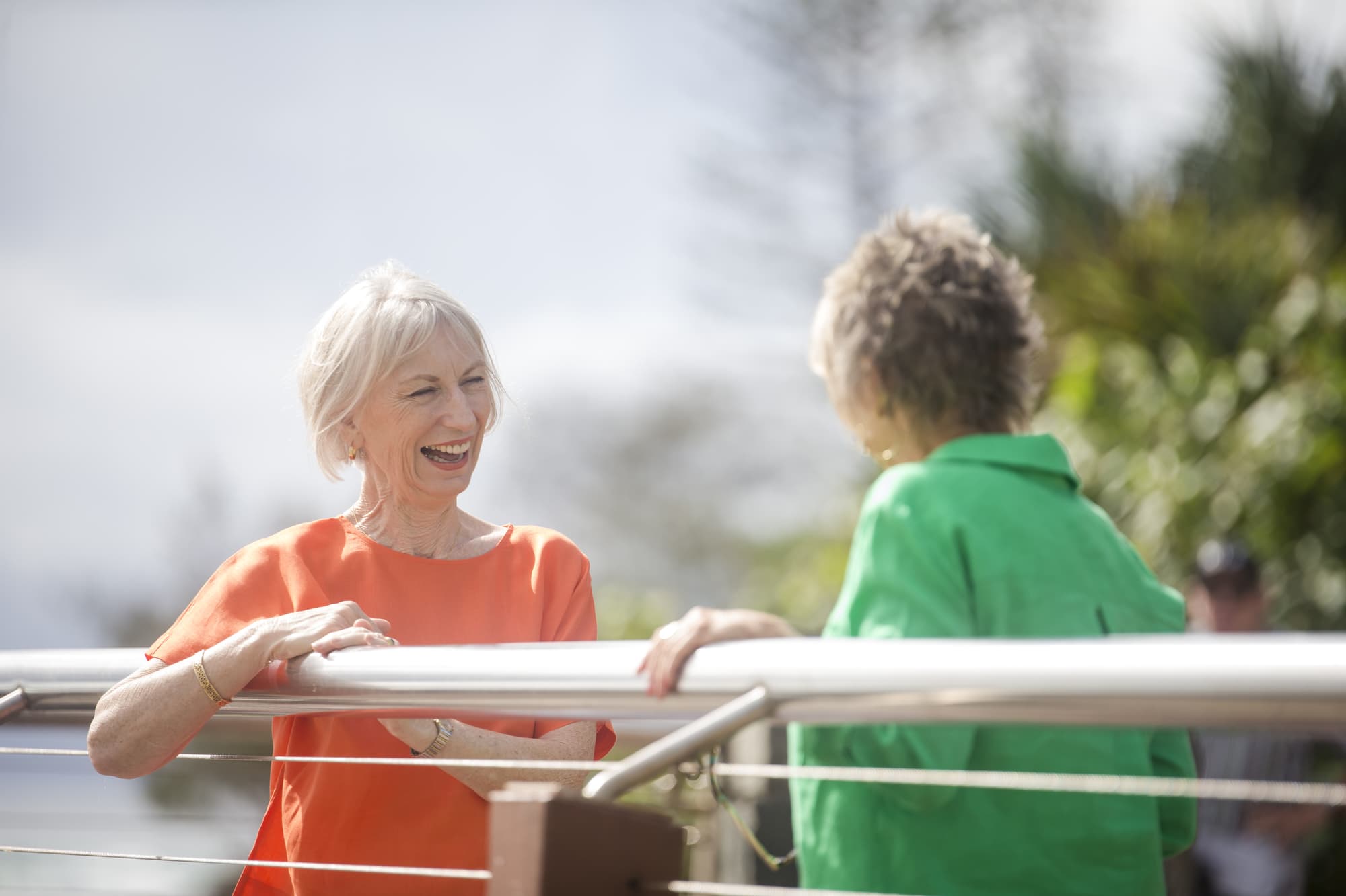 Two older women smiling and talking outdoors near a railing. One wears an orange blouse, the other a green blouse. Trees and a cloudy sky are in the background, creating a relaxed and pleasant atmosphere.