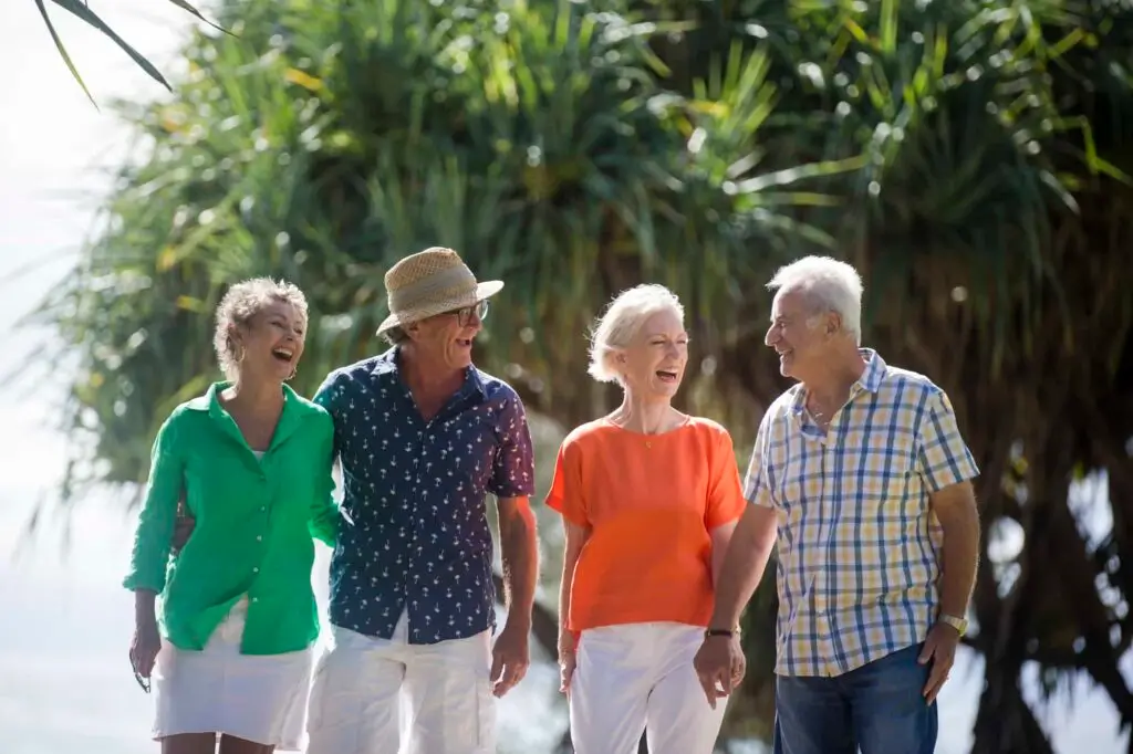 Four older adults, two men and two women, are walking outdoors together at a Sunshine Coast Retirement Community. They are smiling and laughing on a sunny day, with large green tropical plants in the background.