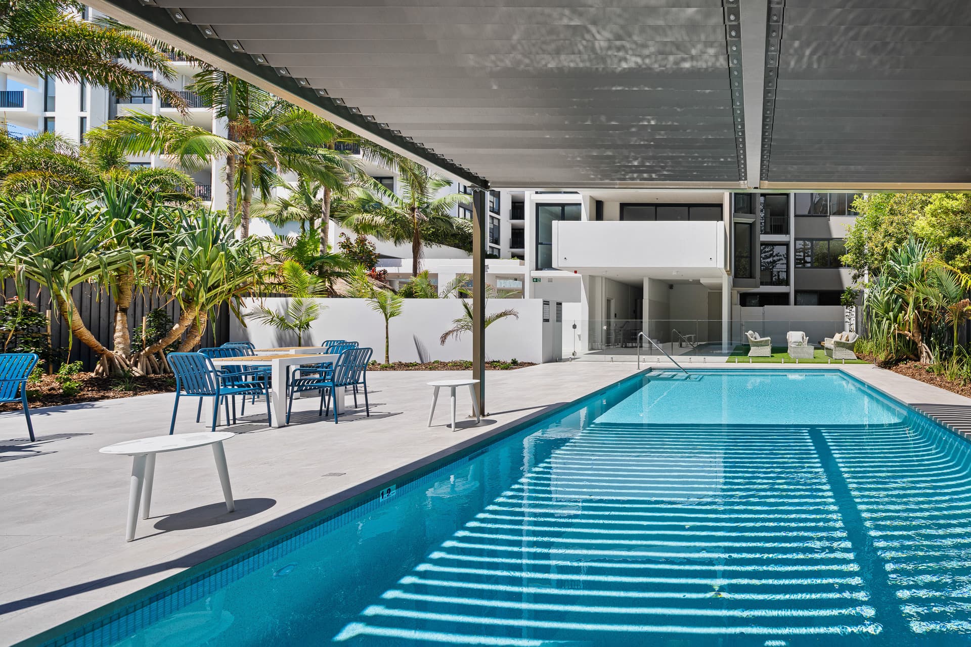 A modern outdoor pool area with clear blue water, surrounded by palm trees and greenery. There are blue chairs and a table under a shaded canopy. White lounge chairs are seen in the background, with a white building nearby.