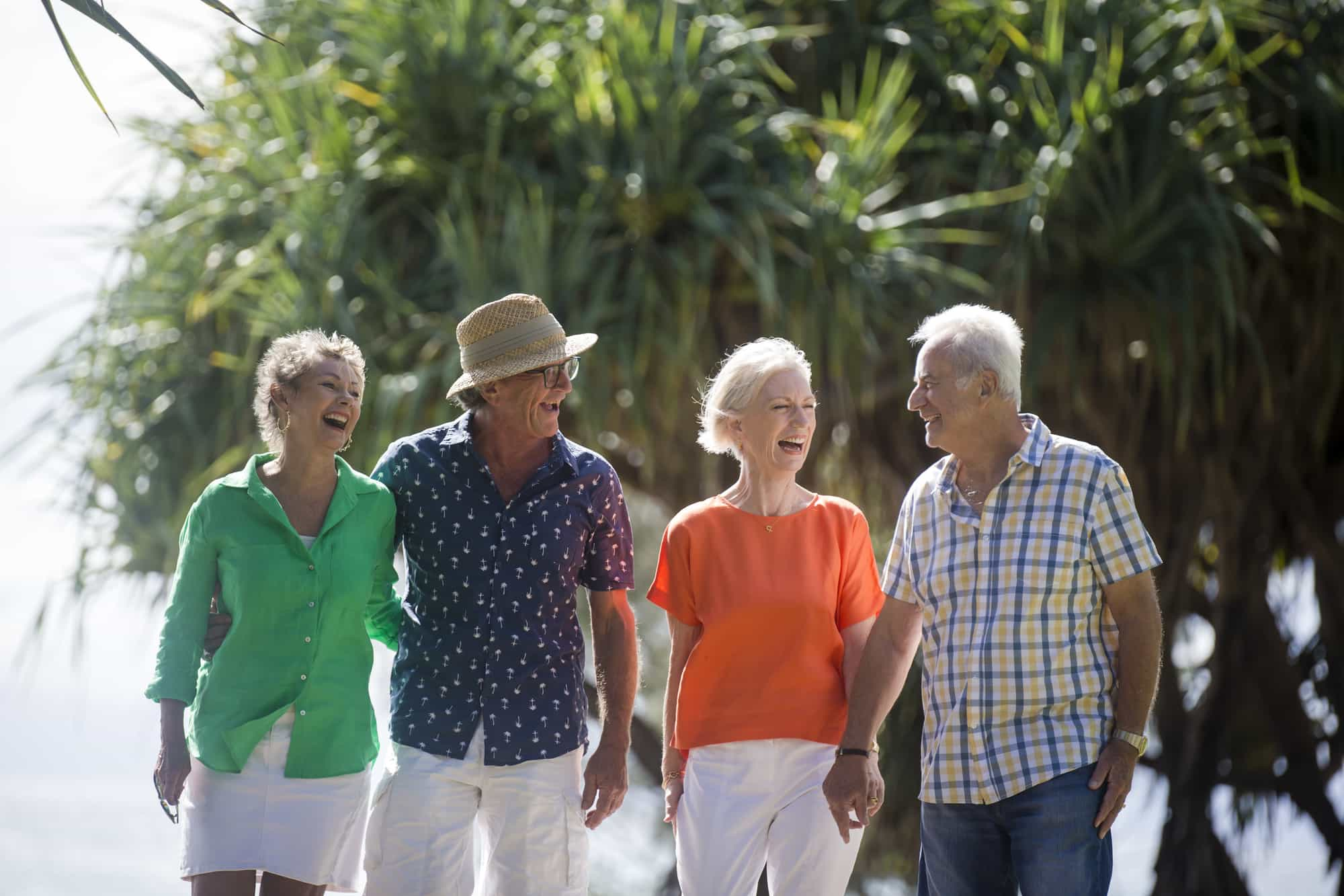 Four older adults are joyfully walking together outdoors. They are dressed in casual, colorful summer clothes. In the background, lush greenery is visible, suggesting a warm, sunny day. All appear to be enjoying each other's company.