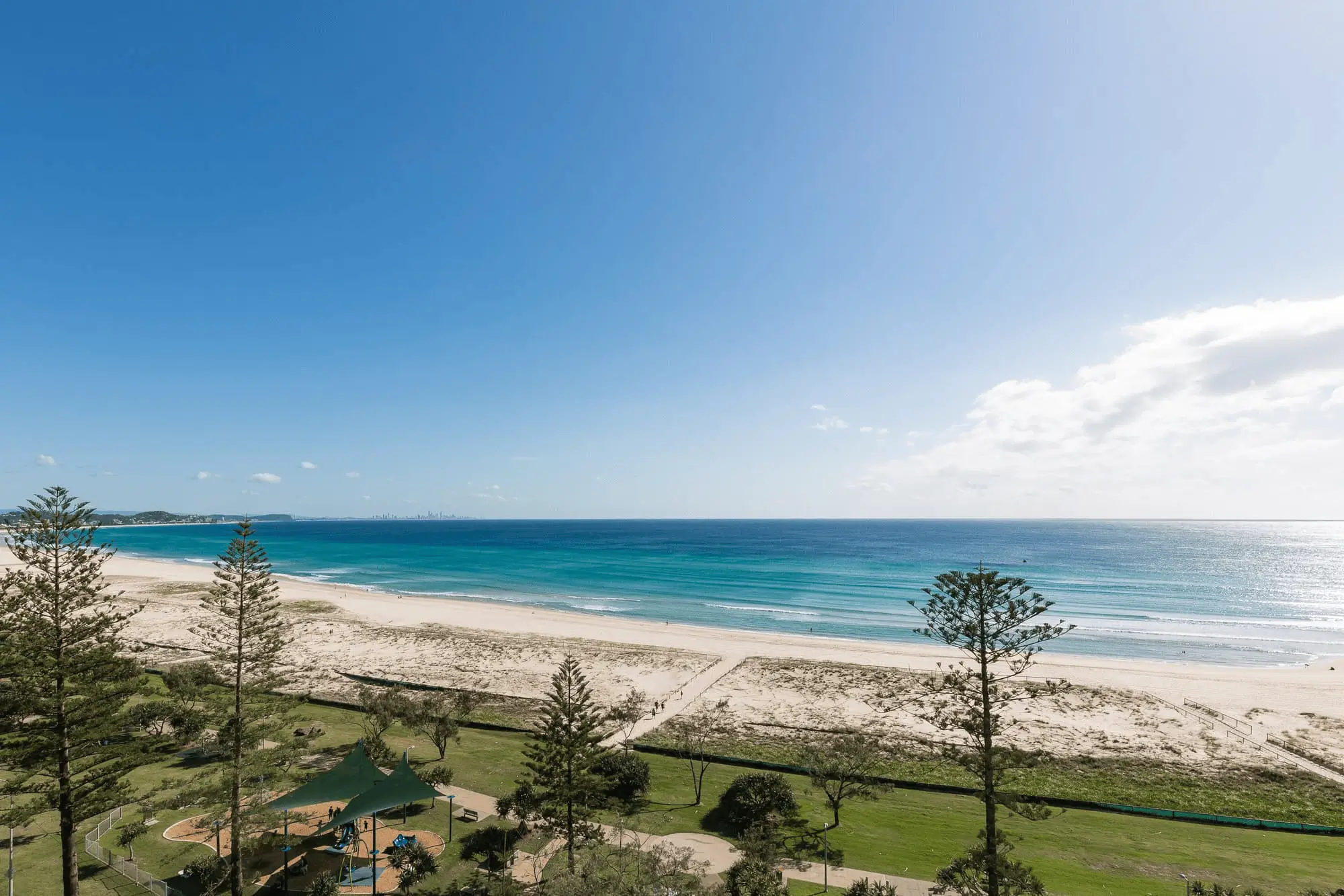 A panoramic view of a sandy beach with turquoise ocean waves, clear blue sky, scattered clouds, and tall pine trees near a grassy park area—perfect for a Gold Coast retirement community.