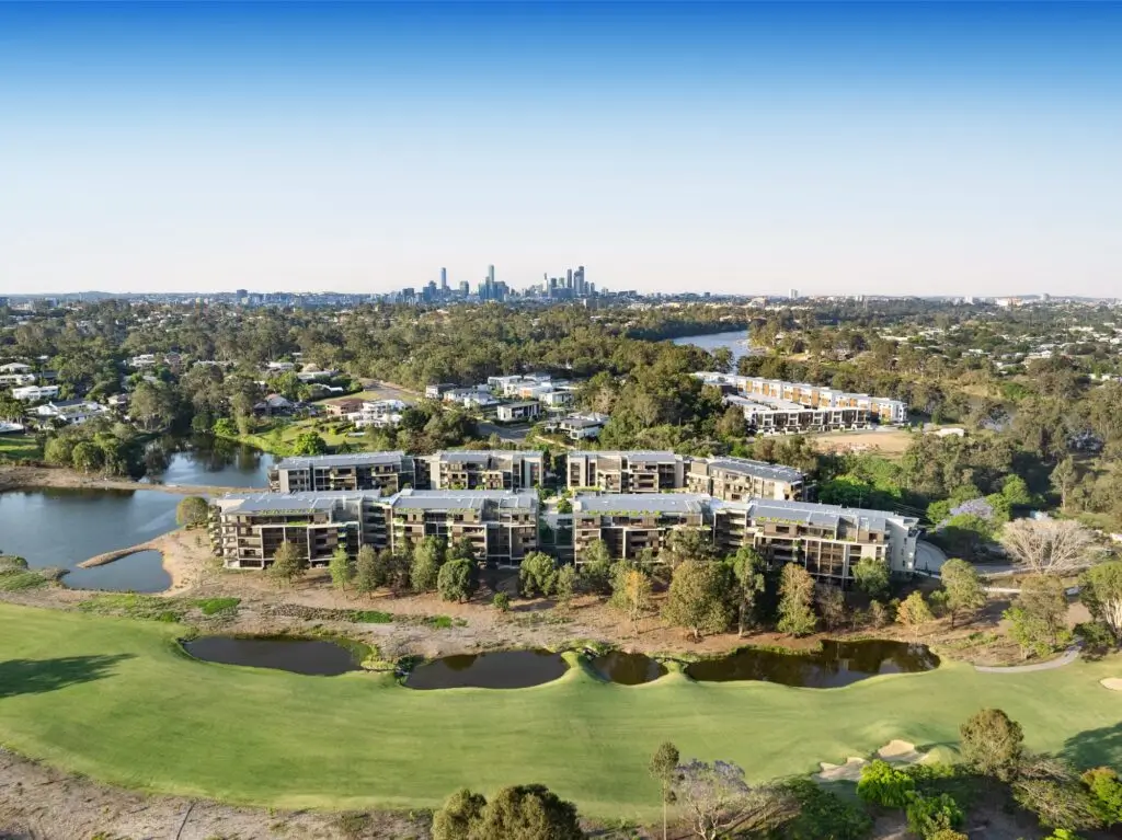 Aerial view of modern apartment buildings in the West Brisbane Retirement Community, surrounded by greenery, small lakes, and a golf course, with a city skyline visible in the distance under a clear blue sky.