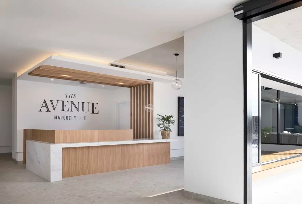 Modern reception area with a wooden and white marble desk, potted plant, and "The Avenue Maroochydore" sign on the wall. Ceiling features wood slats and pendant lights, creating a welcoming entrance to this Sunshine Coast Retirement Community.