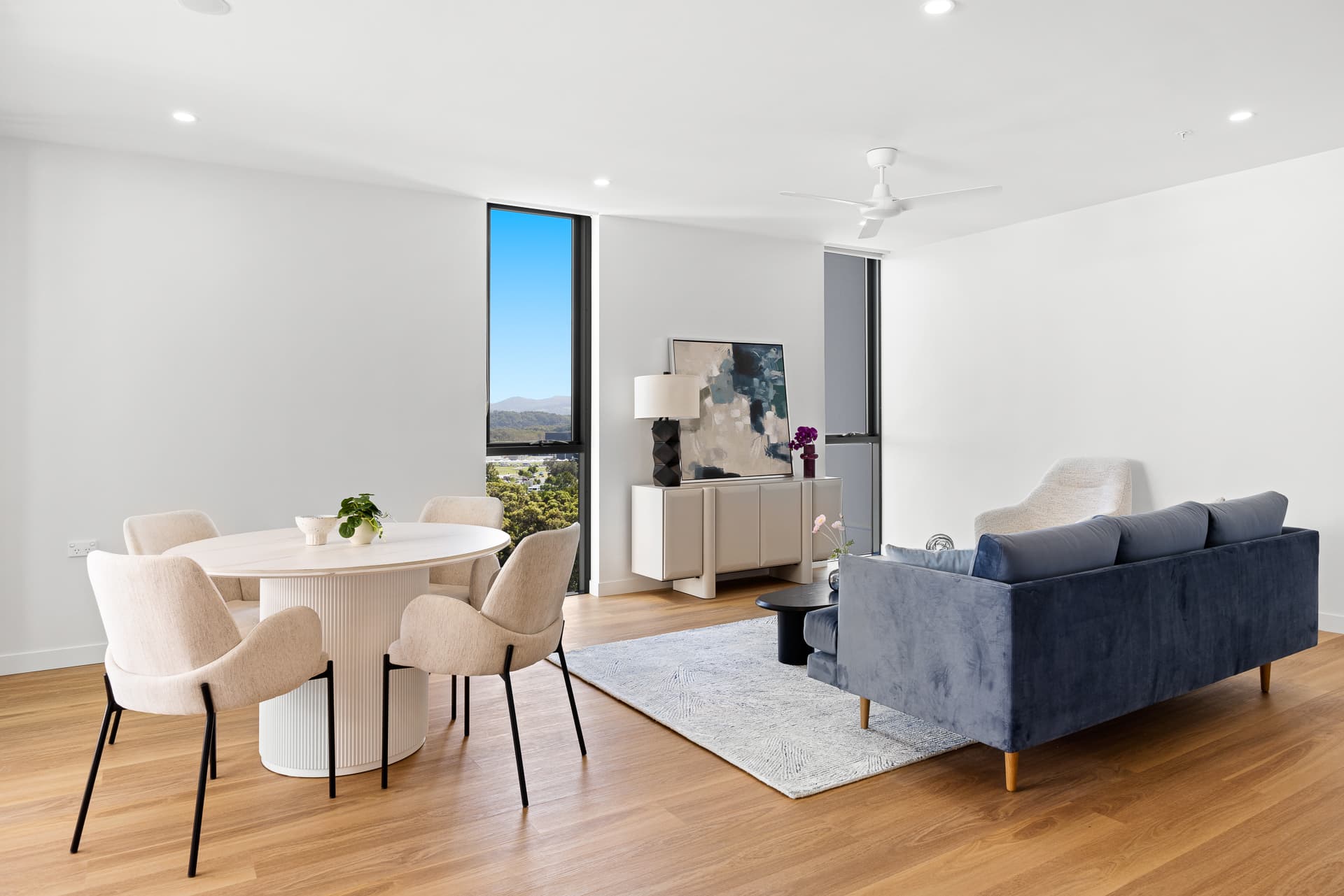 A modern living and dining area with a round white table and beige chairs on a wooden floor, a blue sofa facing a TV, and a large window showcasing a scenic mountain view. Decor includes a rug, plants, and a ceiling fan.