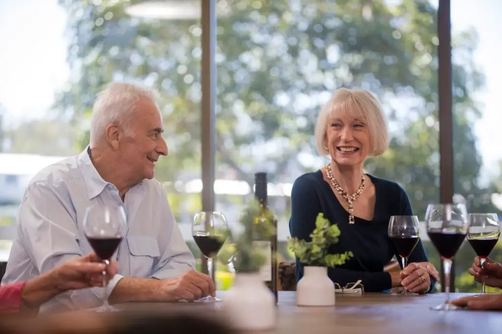 Two older adults sit at a table in the West Brisbane Retirement Community, smiling and enjoying glasses of red wine with friends. Sunlight streams through large windows with trees outside, creating a relaxed, joyful atmosphere.