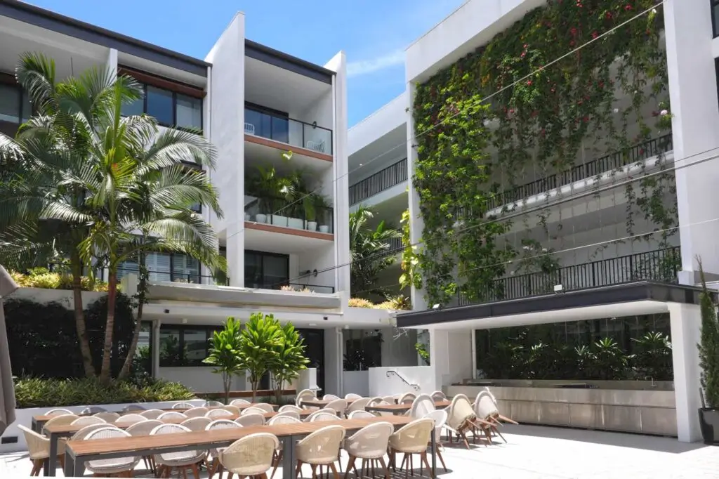 Modern outdoor dining area with long tables and chairs, surrounded by contemporary white buildings with balconies, tropical plants, and a vertical garden wall—perfect for socializing at a Sunshine Coast Retirement Community under a clear blue sky.