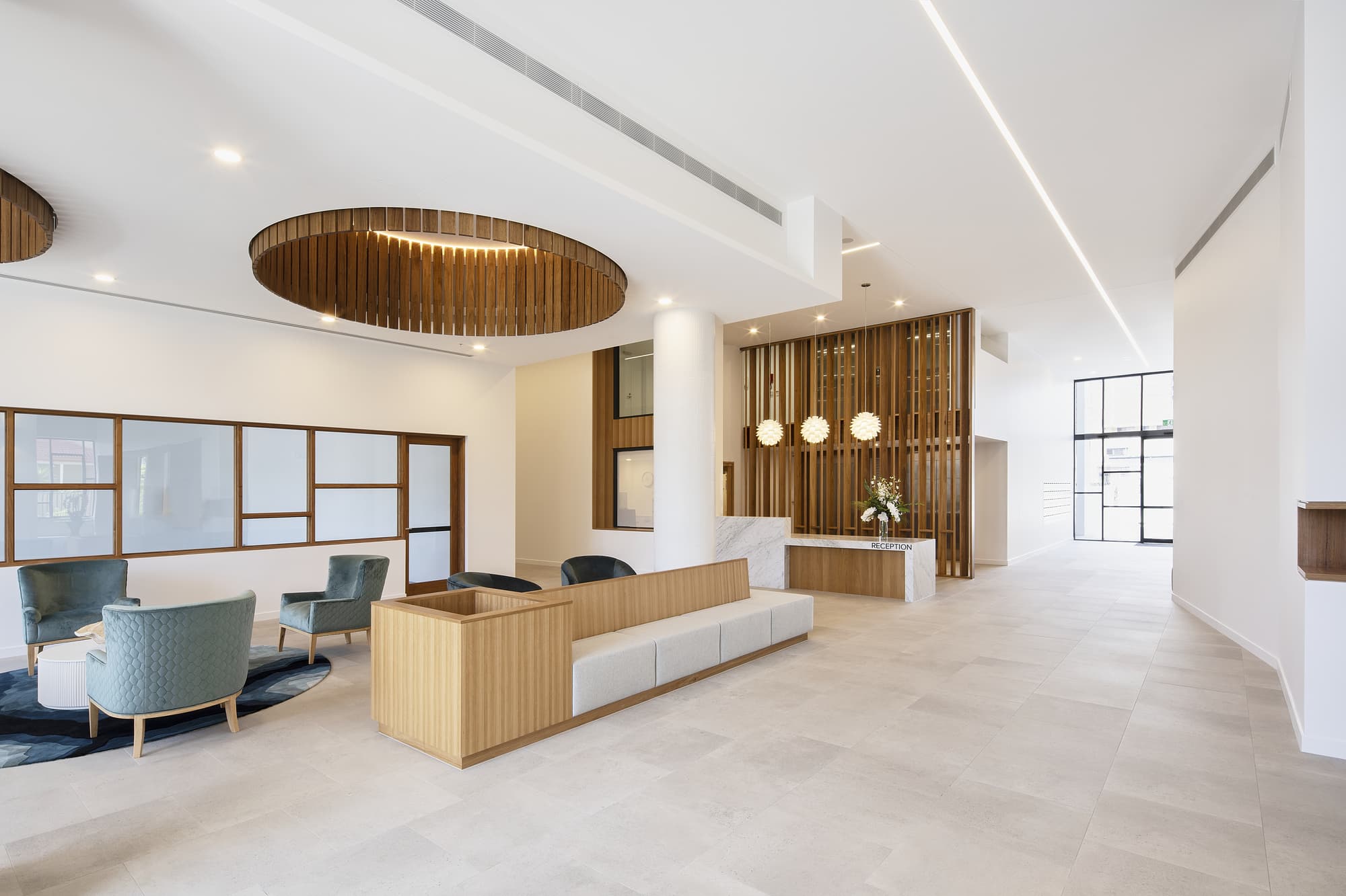 A modern lobby with minimalist design features light stone flooring, wooden panel accents, and large circular ceiling fixtures. There are cushioned chairs, low tables, and a reception desk decorated with hanging lights and floral arrangements.