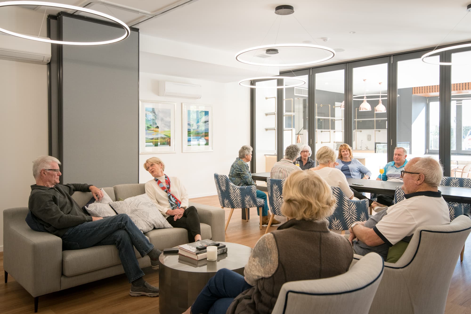 A group of elderly people sit and chat in a modern, well-lit communal living area with sofas and a long table. The room has large windows, decorative lighting, and framed artwork on the walls.