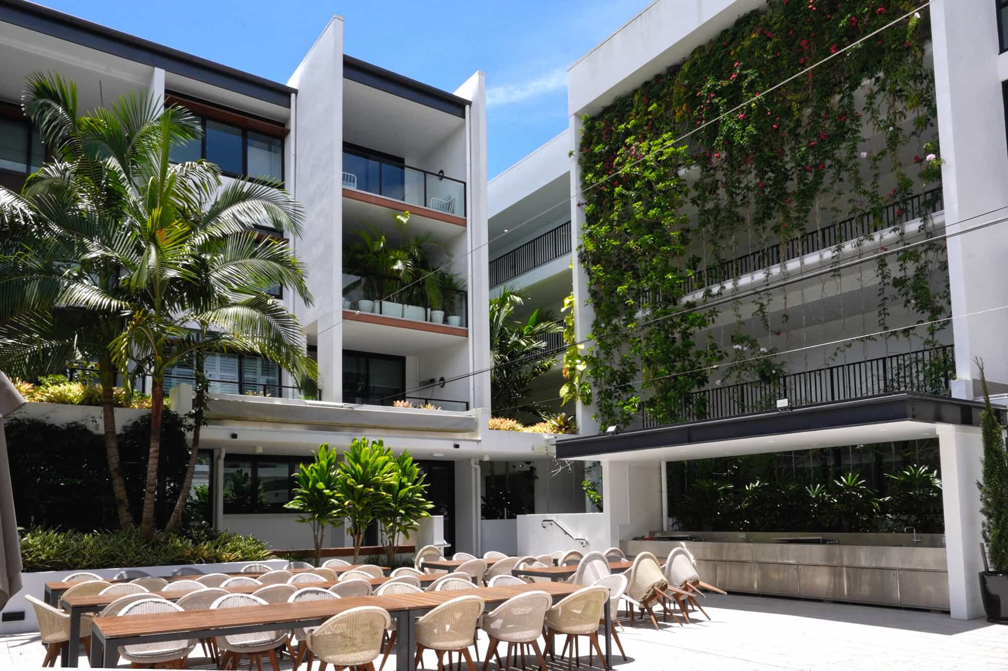 A modern outdoor dining area with a long wooden table and beige chairs is set in front of contemporary white buildings. One building features a lush vertical garden. Palm trees and bright blue sky add to the tropical ambiance.