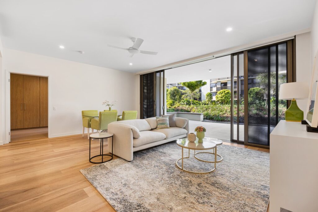 A modern living room with a cream sofa, two round coffee tables, and a carpeted floor. Large glass doors open to a garden view. A dining table with green chairs sits in the corner. Natural light fills the space.