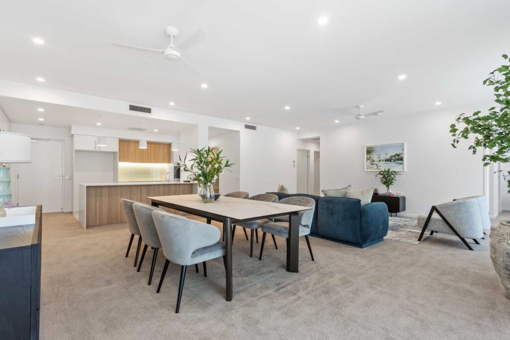 Spacious modern living and dining area with light-colored walls and carpet. Features a wooden dining table with gray chairs, dark sofas, indoor plants, and a kitchen in the background with white cabinets and wooden accents. Ceiling fans installed.