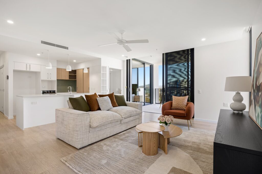 Modern living room with a neutral-toned sofa, cushions, and an armchair. A round wooden coffee table holds flowers. The open-plan space includes a kitchen with white cabinets and hanging lights. Large glass doors open to a balcony with a view.