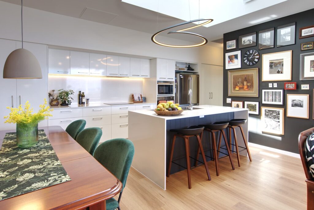 Modern kitchen and dining area with white cabinets and a central island featuring fruit bowls and wooden stools. The wall displays framed pictures and a clock. A wooden dining table and green chairs sit to the left, with a vase of yellow flowers on top.