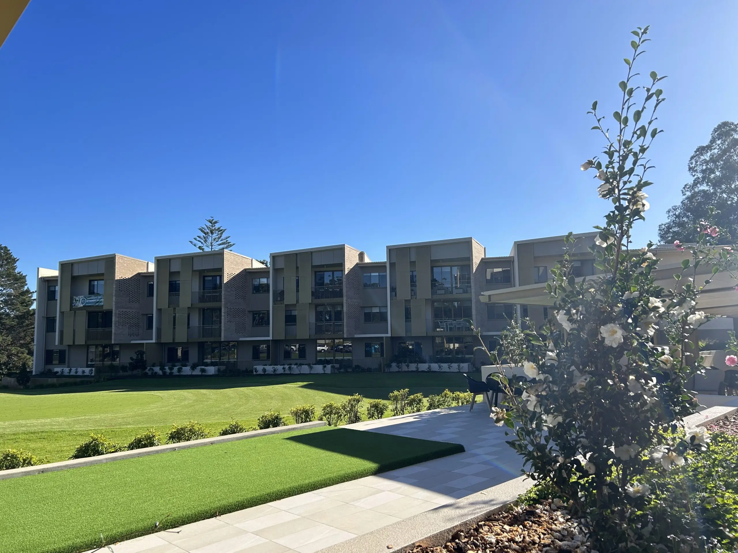 A modern, multi-story building with large windows overlooks a manicured lawn under a bright blue sky at the Toowoomba Retirement Community. Flowering shrubs and landscaped greenery line the foreground.
