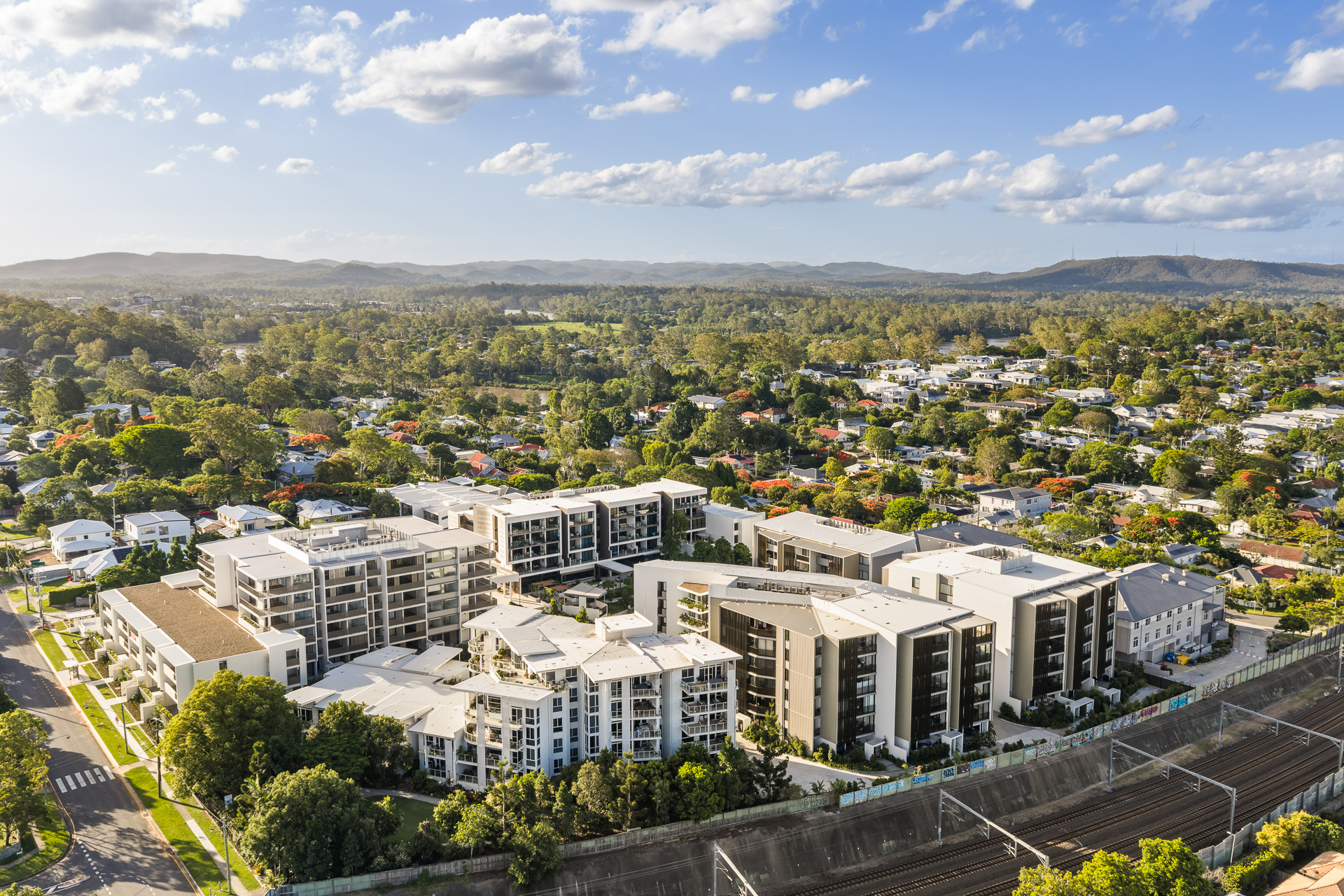 Aerial view of a modern Brisbane Retirement Community apartment complex surrounded by trees and suburban houses, with hills and greenery visible in the background under a partly cloudy sky.