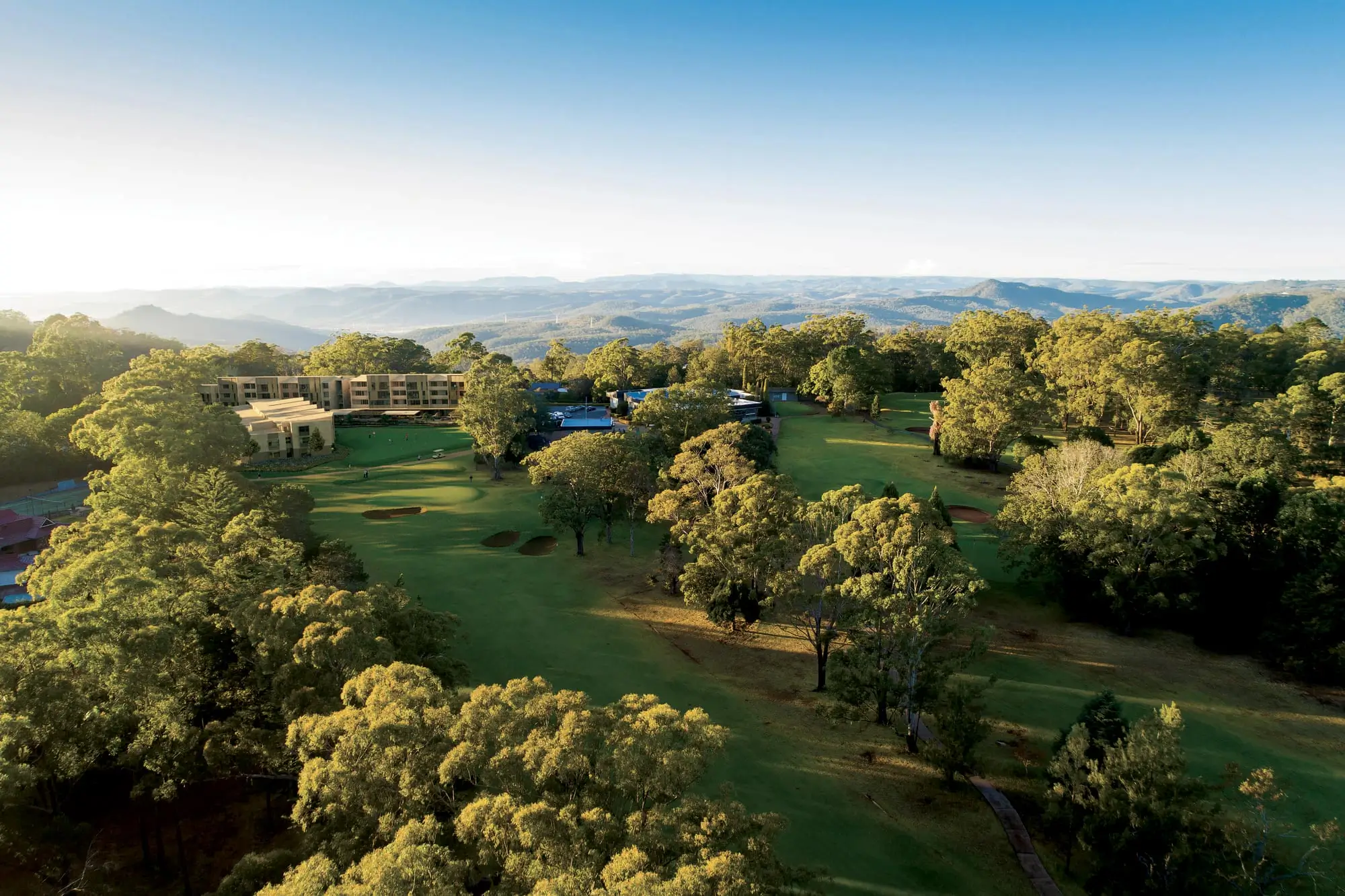 Aerial view of a golf course surrounded by dense trees, with buildings on the left from the nearby Toowoomba Retirement Community and mountains in the distant background under a clear blue sky.