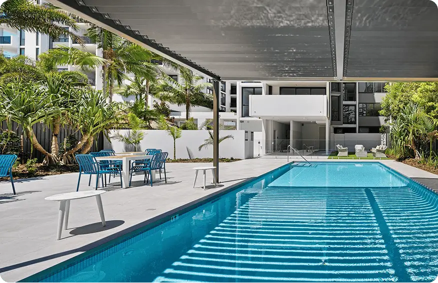 Outdoor swimming pool next to a modern apartment building in a Gold Coast retirement community, with blue chairs and tables under a pergola, surrounded by palm trees, plants, and lounge chairs in the background.