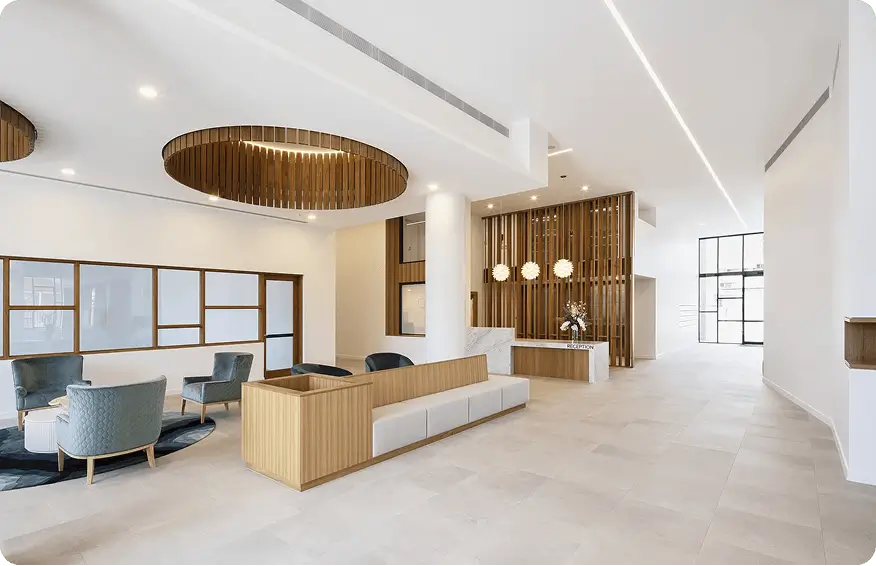 A modern, spacious Gold Coast retirement community lobby features neutral tones, wood accents, a seating area with blue chairs, white sofas, circular ceiling lights, and large windows letting in natural light.