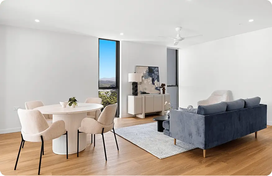 Modern living and dining area with light wood flooring, a round dining table with four beige chairs, a blue sofa, a white sideboard, and a large window with scenic views—perfect for relaxed days in this Gold Coast retirement community. Decor is minimal and contemporary.