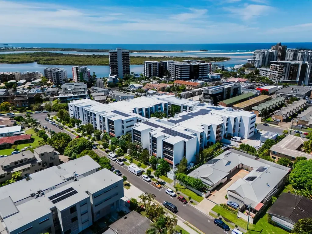 Aerial view of a modern coastal city with white apartment buildings, houses, roads, greenery, and waterways—ideal for a Sunshine Coast Retirement Community—with high-rises and the ocean in the background under a blue sky.