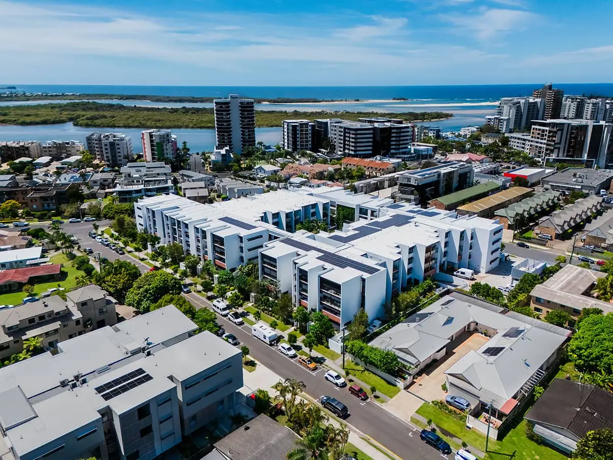 Aerial view of a modern coastal city with white apartment buildings, houses, roads, greenery, and waterways—ideal for a Sunshine Coast Retirement Community—with high-rises and the ocean in the background under a blue sky.