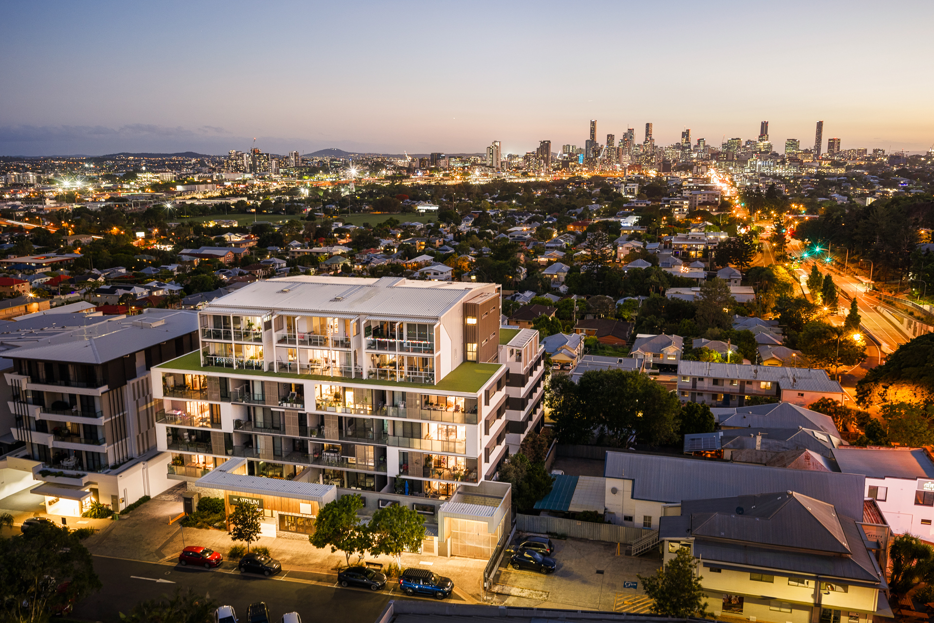 Aerial view of a residential neighborhood at dusk, with The Atrium—a modern mid-rise apartment building—in the foreground and a brightly lit city skyline in the background. Streets and houses are illuminated by streetlights.
