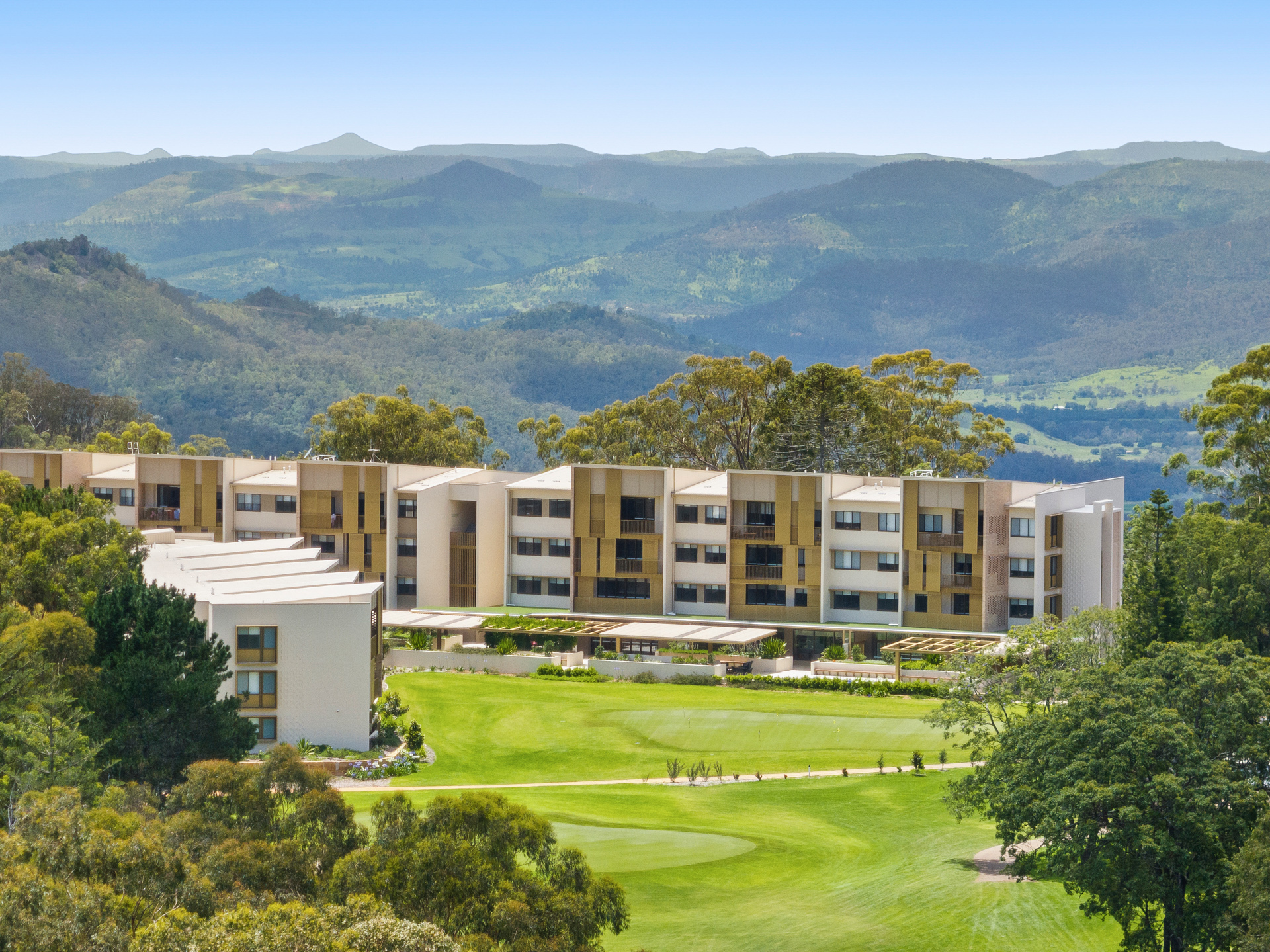 A modern building complex with multiple levels is surrounded by lush greenery and a golf course. In the background, rolling hills and a clear sky create a serene landscape.