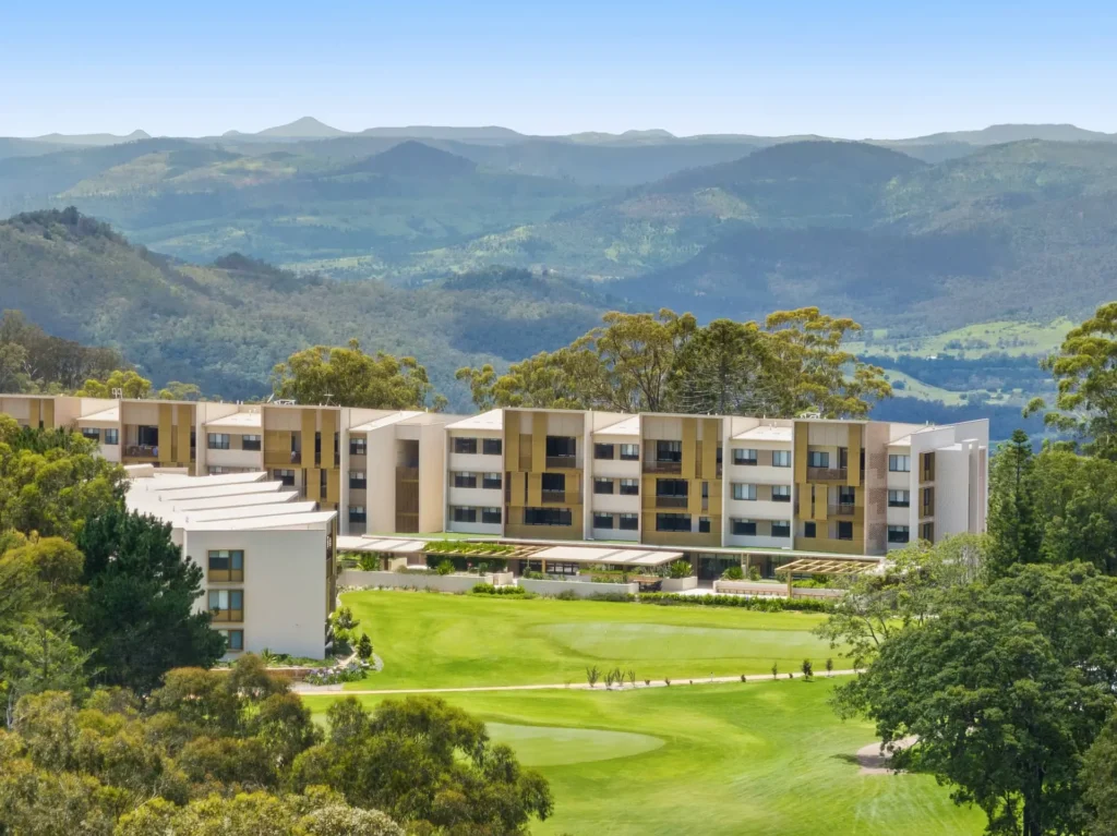 A modern apartment complex with beige and white facades sits amidst green lawns and tall trees in the serene Toowoomba Retirement Community, with forested hills and blue mountains in the background under a clear sky.