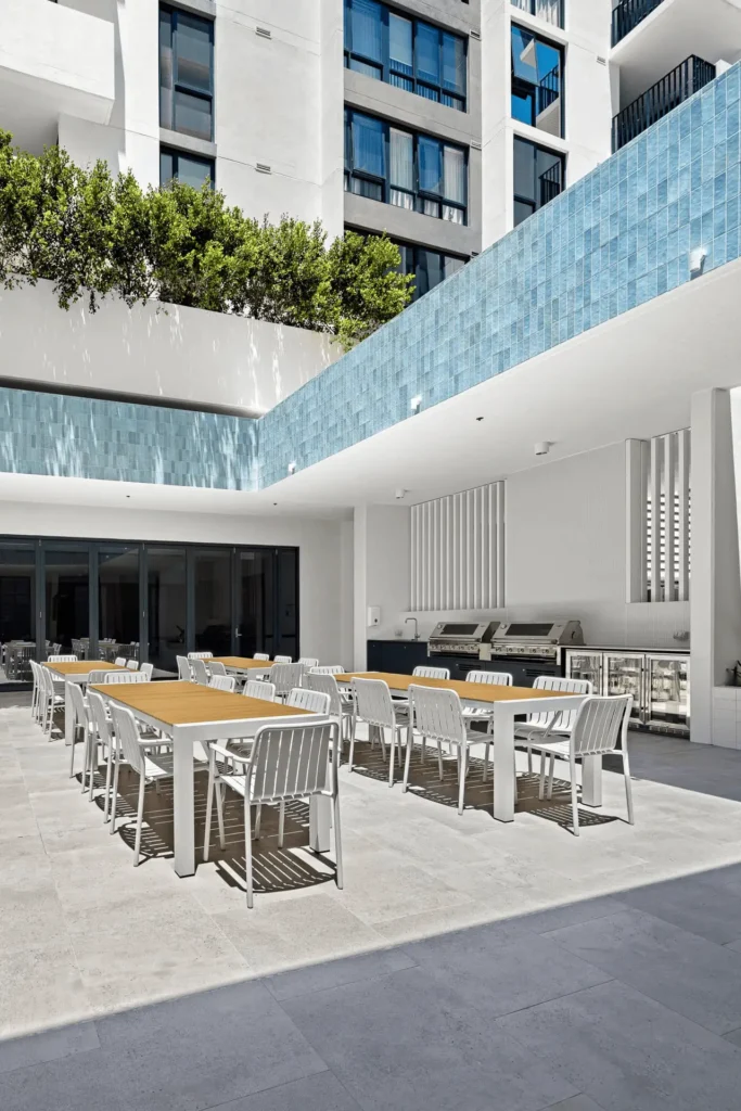 Modern outdoor dining area with wooden tables and white chairs, set under a blue-tiled overhang near a building in a Gold Coast retirement community. Built-in grills and greenery are visible, with sunlight casting shadows on the patio.