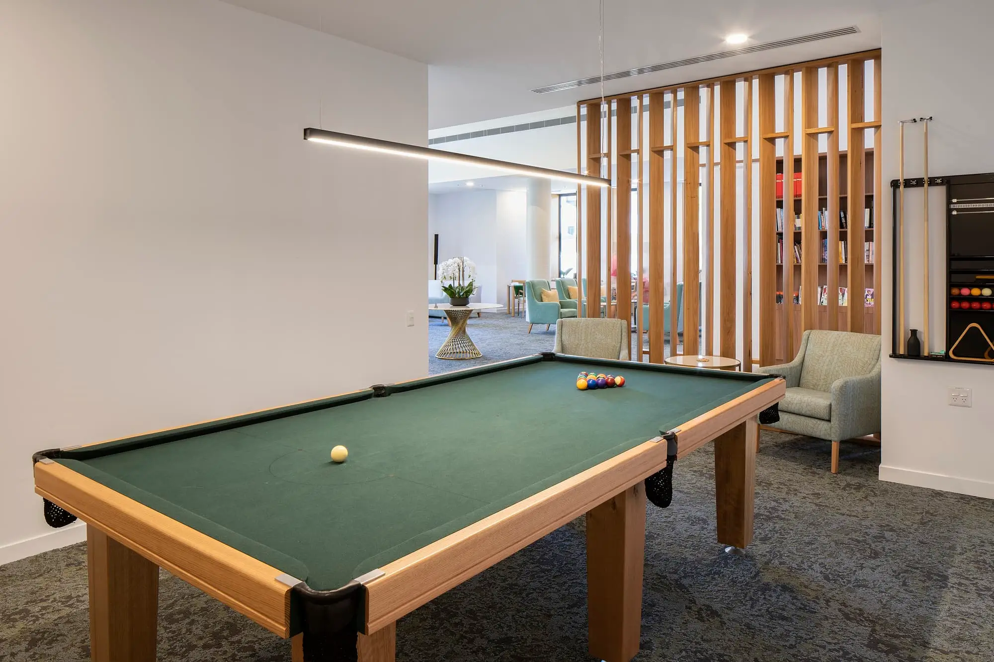 A modern room in this Gold Coast retirement community features a pool table with green felt, balls racked for play, a cue stick holder on the wall, lounge chairs, and a wooden slat divider separating it from a seating area.