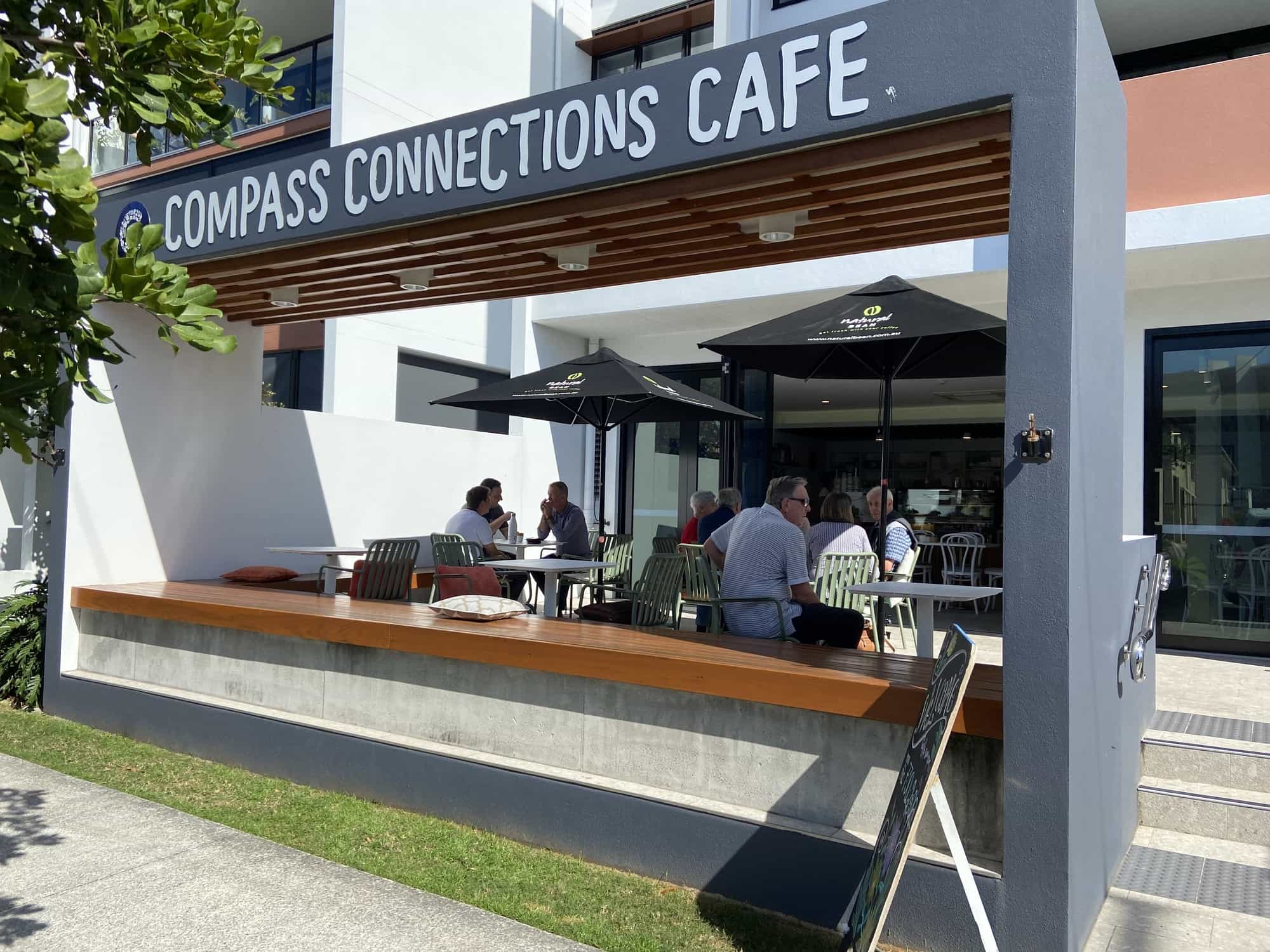 Outdoor seating area at Compass Connections Cafe, a popular spot in the Sunshine Coast Retirement Community. Several people sit under large black umbrellas. The cafe features a modern grey and white exterior with a wooden bench along the front.