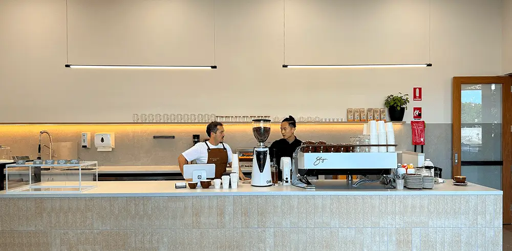 Two baristas stand behind a modern, minimalist coffee bar in a Gold Coast retirement community, with coffee machines, cups, and pastries on display. The counter is clean and well-lit, and shelves with glasses line the wall behind them.