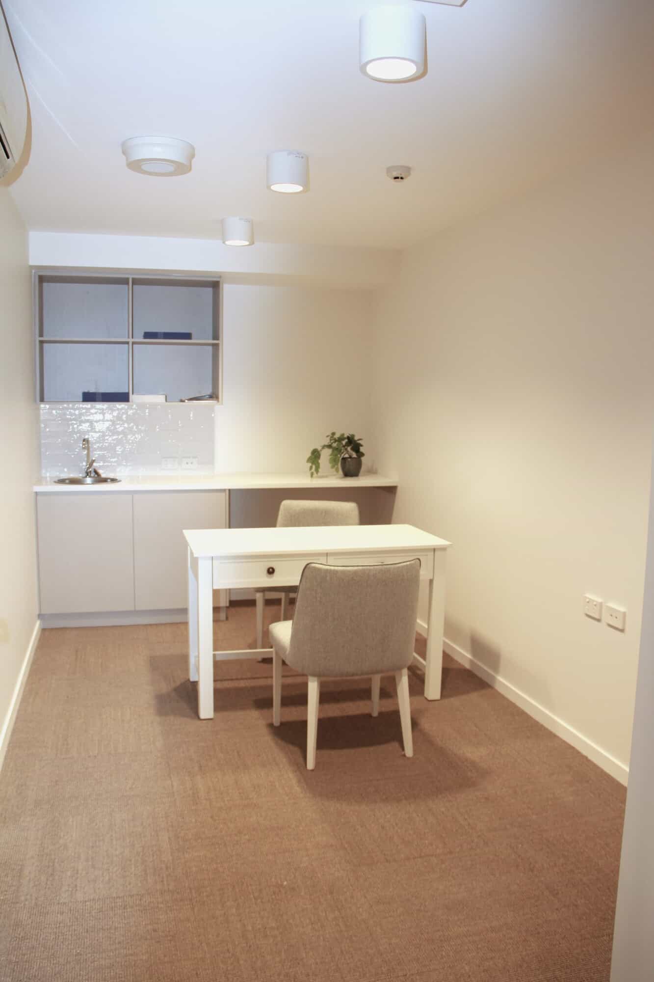 A small, minimalist office with white walls, a desk, and two beige chairs—perfect for consultations in a Sunshine Coast Retirement Community. A compact kitchenette and potted plant add warmth to this well-lit, inviting space.